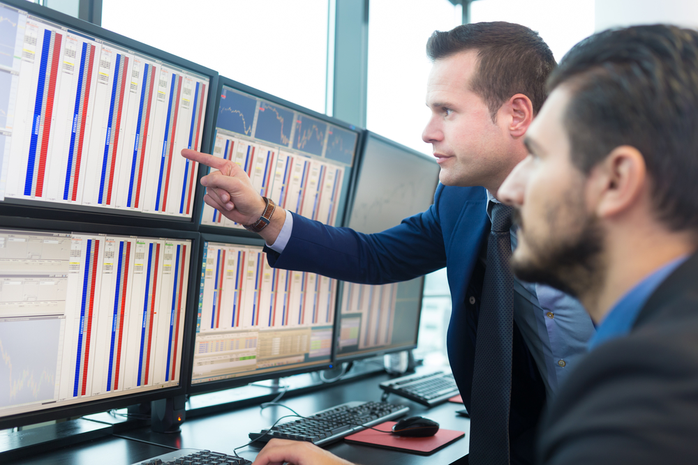 Businessmen trading stocks. Stock traders looking at graphs, indexes and numbers on multiple computer screens. Colleagues in discussion in traders office.