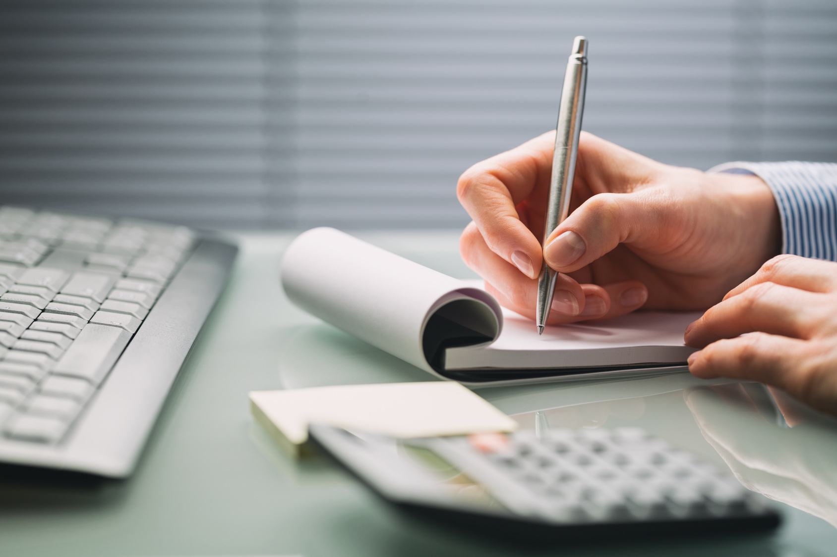 A female hand takes notes on a paper notebook over a busy office desktop. Low angle closeup image.