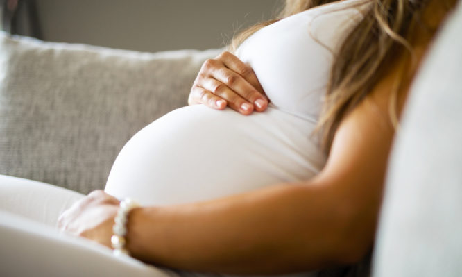 Close-up of pregnant woman relaxing and sitting on the side on the sofa. Holding a hands on the tummy.