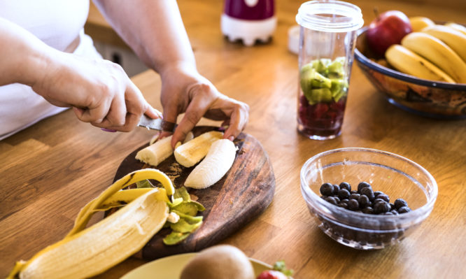 Unrecognizable overweight woman in white t-shirt at home cutting a banana for a delicious healthy smoothie in her kitchen.