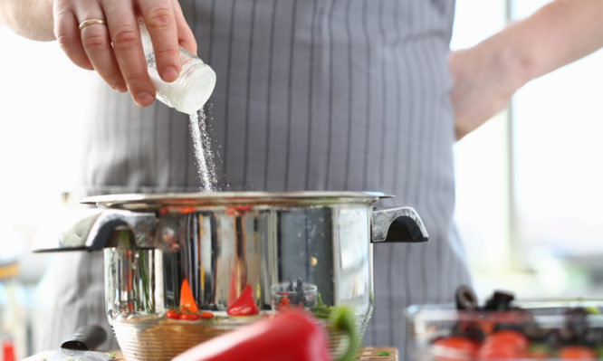 Culinary Chef Adding Saucepan White Sea Salt. Man in Apron Holding Spice Shaker in Hand. Male Fingers Putting Ingredient to Stainless Pan. Cooking in Kitchen at Home Horizontal Photography