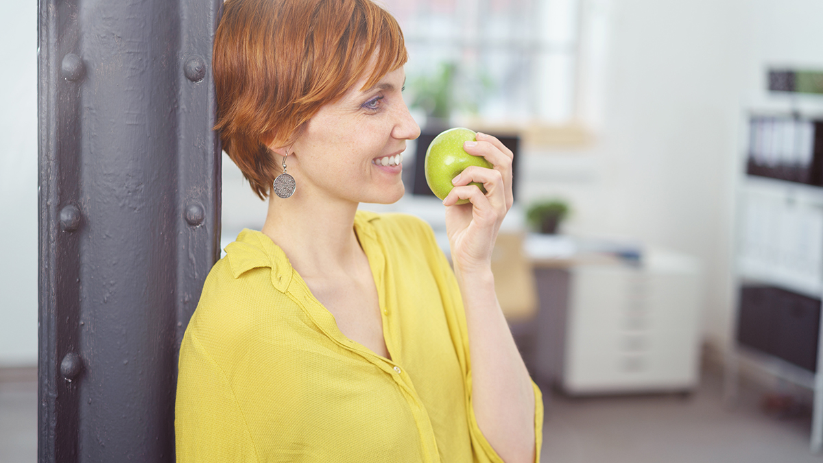 Profile view on pretty young red haired woman in yellow blouse standing up near post in small office eating a green apple