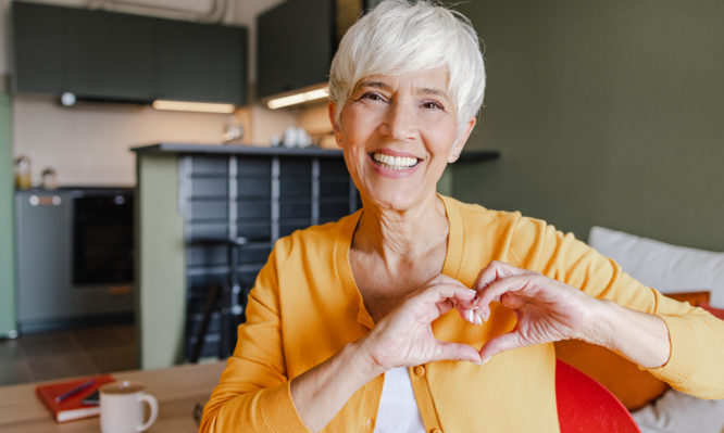 Senior woman is at home, she is in the living room and showing a heart-shaped symbol