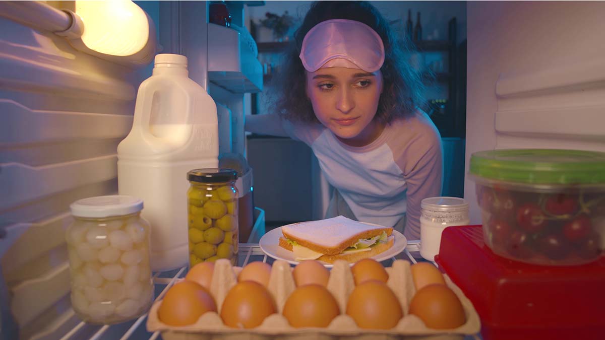 Young sleepy woman opens door of fridge thinking about what to eat at night time. View from fridge