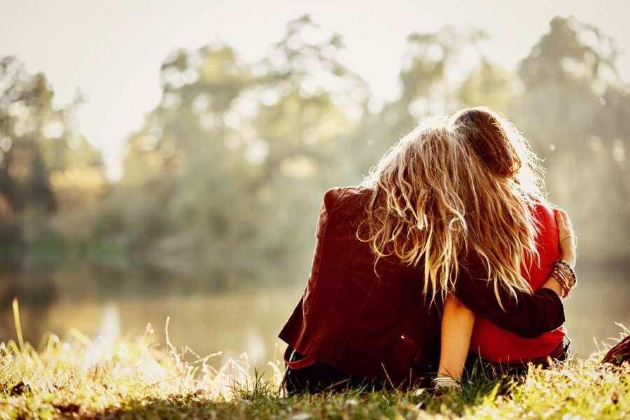 two young women sitting on grass hugging rear view