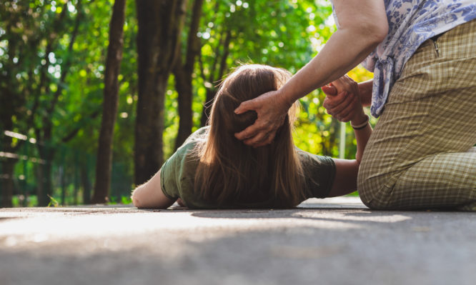 Fainted girl helped by an old woman – Teenager trying to get back on her feet while receiving support from an elder