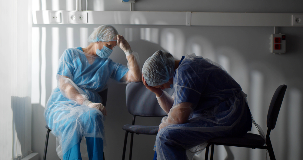 Tired surgeons in sterile gown and safety mask sitting on chairs in hospital room and resting. Portrait of exhausted medical workers in protective uniform relaxing in clinic corridor