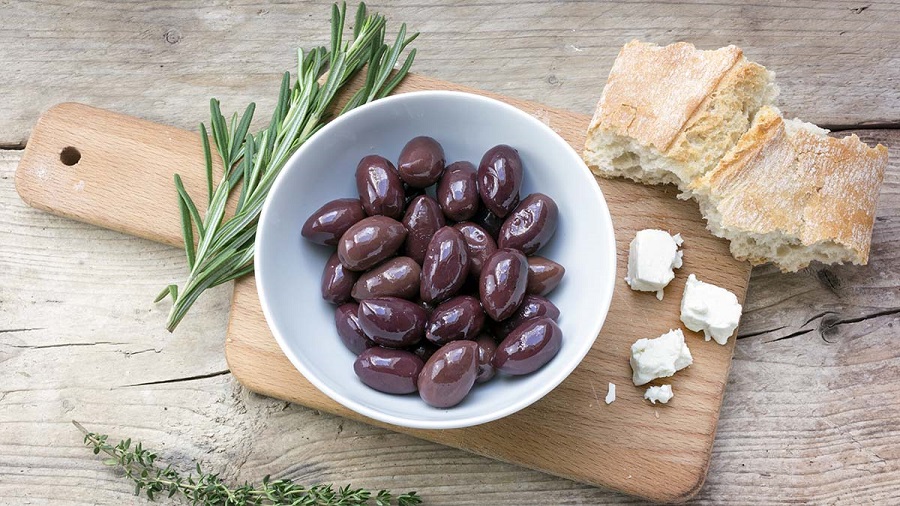 Kalamata black olives in a white bowl, bread, feta cheese and herbs garnish on a rustic wooden table, view from above
