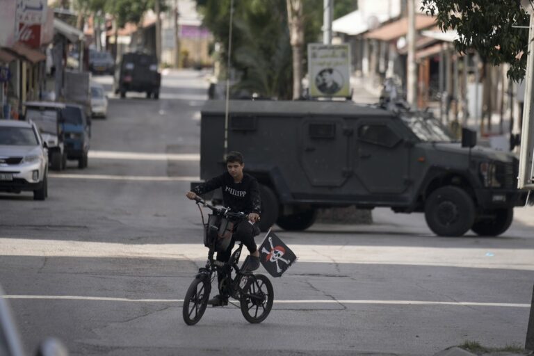 A youth rides his bicycle past an Israeli armoured vehicle during an army raid, following a car that was hit by bullets in which a Palestinian was reportedly killed by Israeli special force, in the occupied West Bank city of Qalqilya, Thursday, Dec. 12, 2024.(AP Photo/Majdi Mohammed).