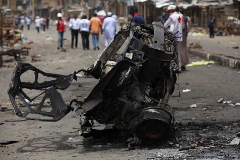 People walk past the remains of one of Tuesday's car bombs in Jos, Nigeria, Wednesday, May 21, 2014. Two car bombs exploded at a bustling bus terminal and market the central Nigeria city, killing at least 118 people, wounding dozens and leaving bloodied bodies amid the flaming debris.(AP Photo/Sunday Alamba)