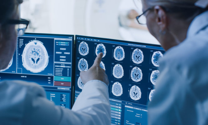 In Control Room Doctor and Radiologist Discuss Diagnosis while Watching Procedure and Monitors Showing Brain Scans Results, In the Background Patient Undergoes MRI or CT Scan Procedure.